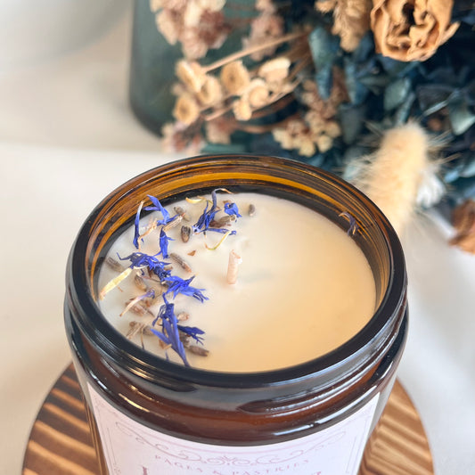 Candle in a glass jar with dried flowers on top, placed on a wooden surface with a blurred background.