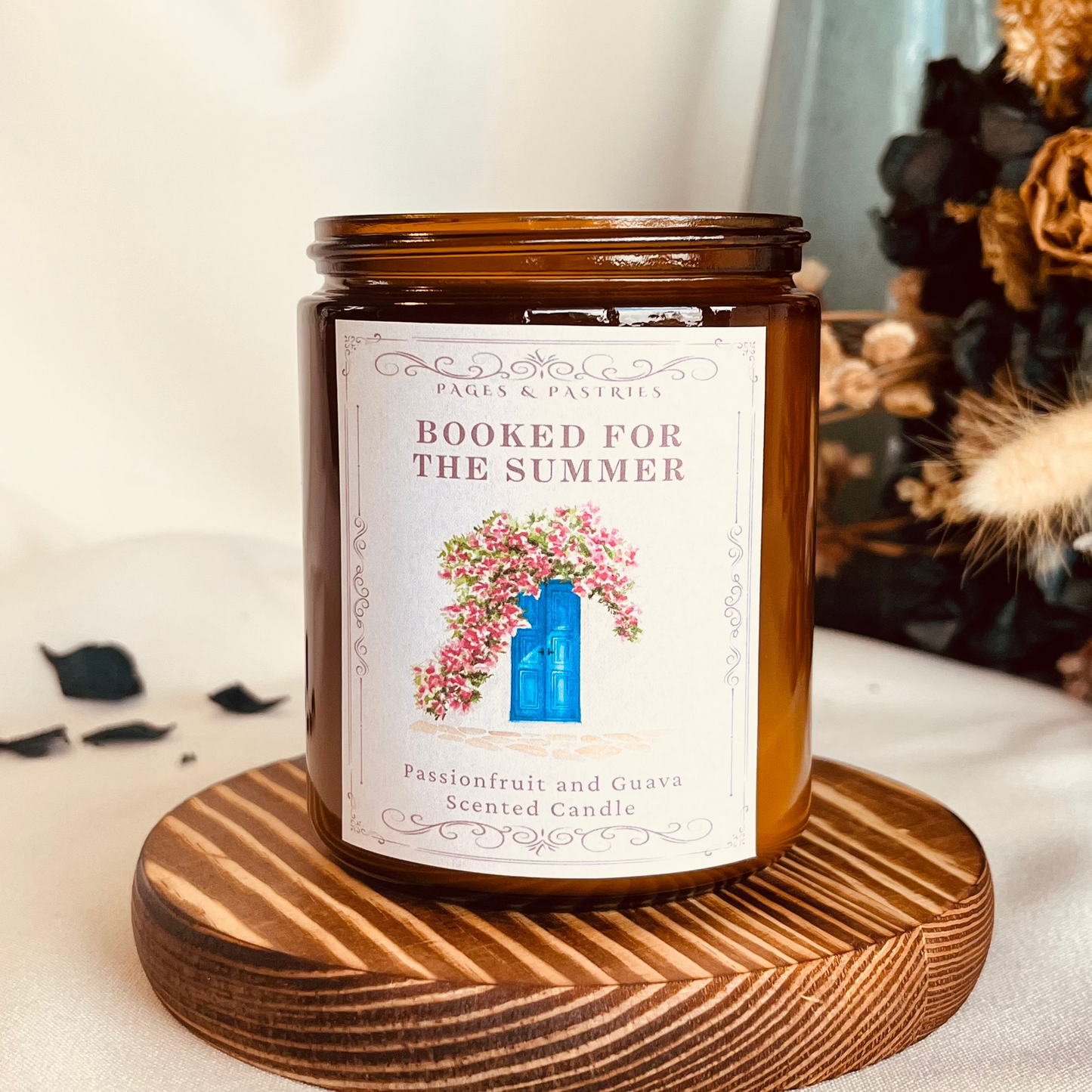 Booked for the Summer bookish candle in amber glass jar with guava and passionfruit scent, featuring blue Greek door with pink hanging flowers on label