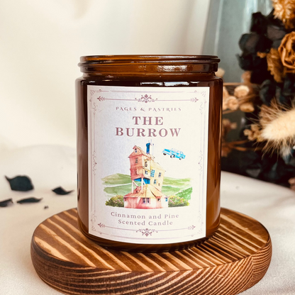 The Burrow bookish candle in amber glass jar with cinnamon and pine scent, featuring label with the Weasley home and flying car