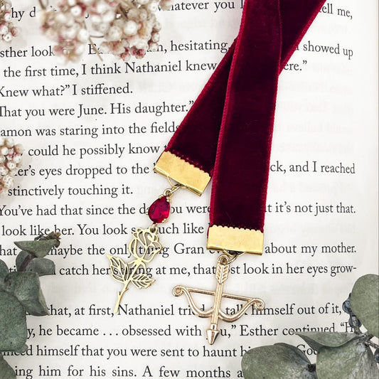A red velvet bookmark with a red zircona and a golden rose on one end and a golden bow and arrow on the other